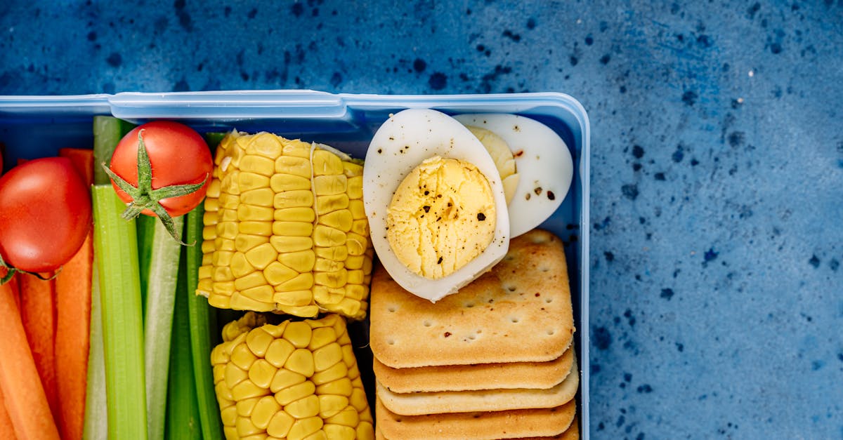 A colorful lunchbox featuring boiled eggs, corn, crackers, and fresh vegetables on a blue background.
